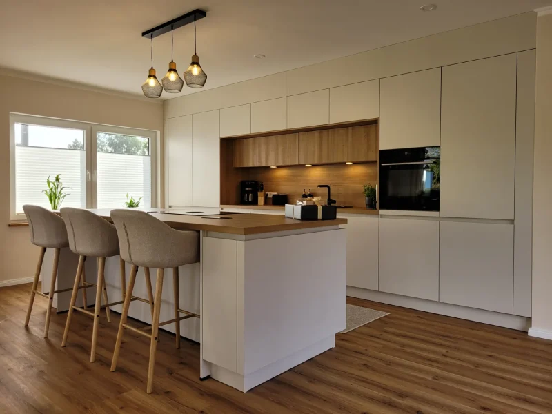 Weiße Designküchenzeile mit Kücheninsel und Barhockern. Die weißen, glatten Fronten werden um Holzelemente ergänzt